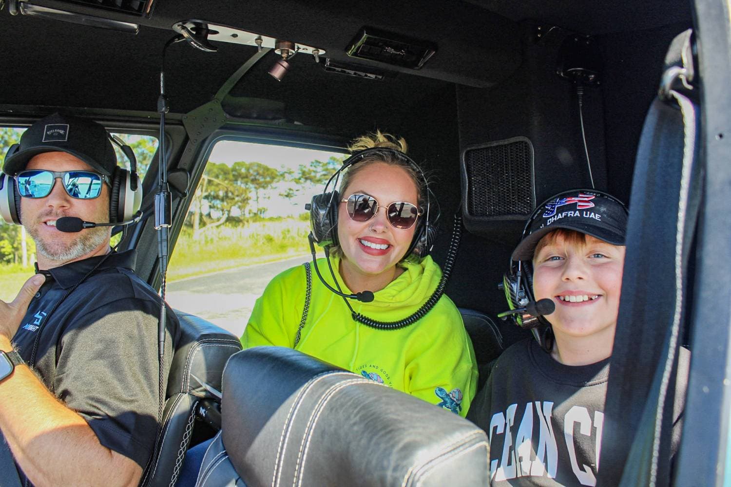 Three people wearing headsets sit in a helicopter cockpit, smiling at the camera. The pilot is on the left, a woman in a neon hoodie sits center, and a boy in a cap is on the right.