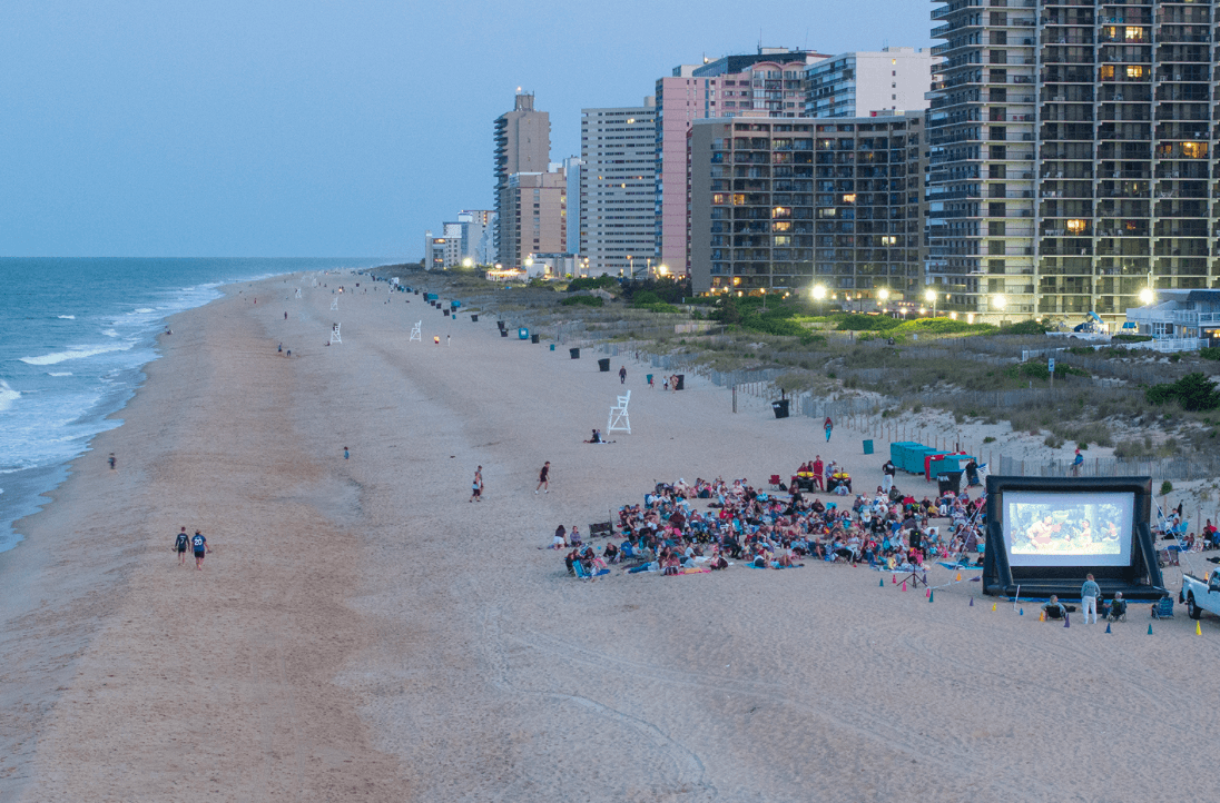 Crowd on the sandy beach watching an outdoor movie projected on a large inflatable screen near beachfront high-rise buildings at dusk.