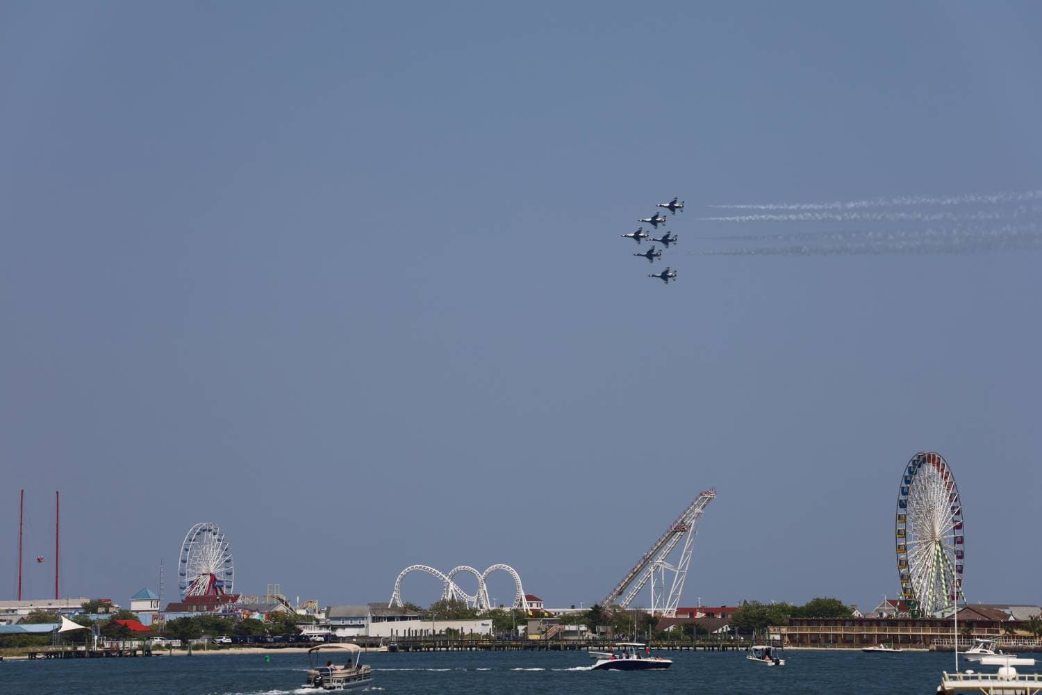 Five military jets fly in tight formation over a coastal amusement park with a ferris wheel and roller coasters, leaving white contrails across a clear blue sky.