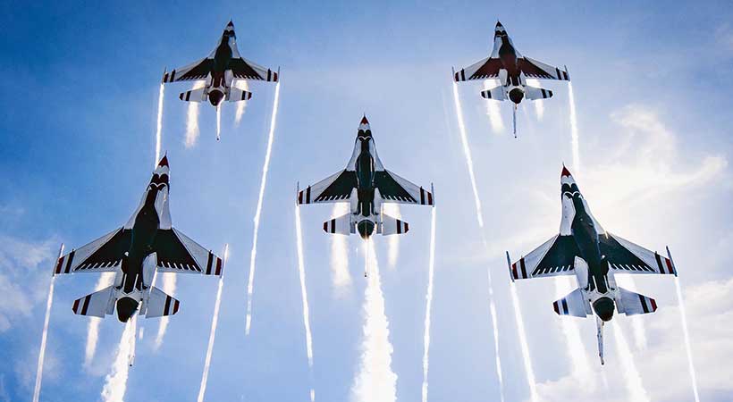 Five fighter jets in a tight formation with smoke trails against a clear blue sky across an air show.